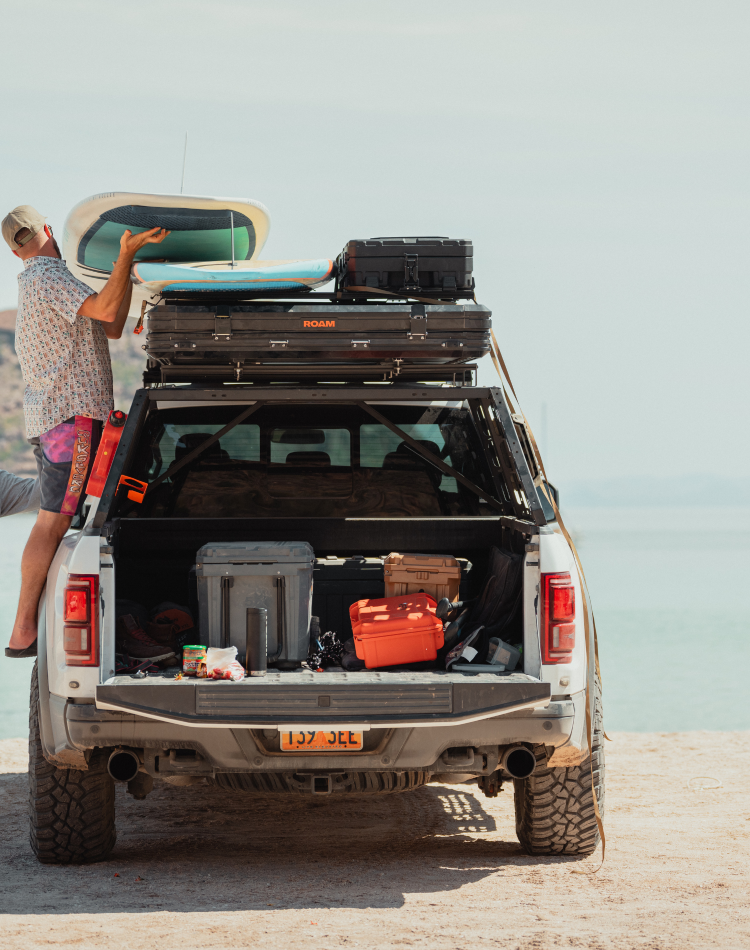 A man lifting a paddle board onto the top of his car while parked at the beach with the Roadie first aid kit and other gear in his trunk. 
