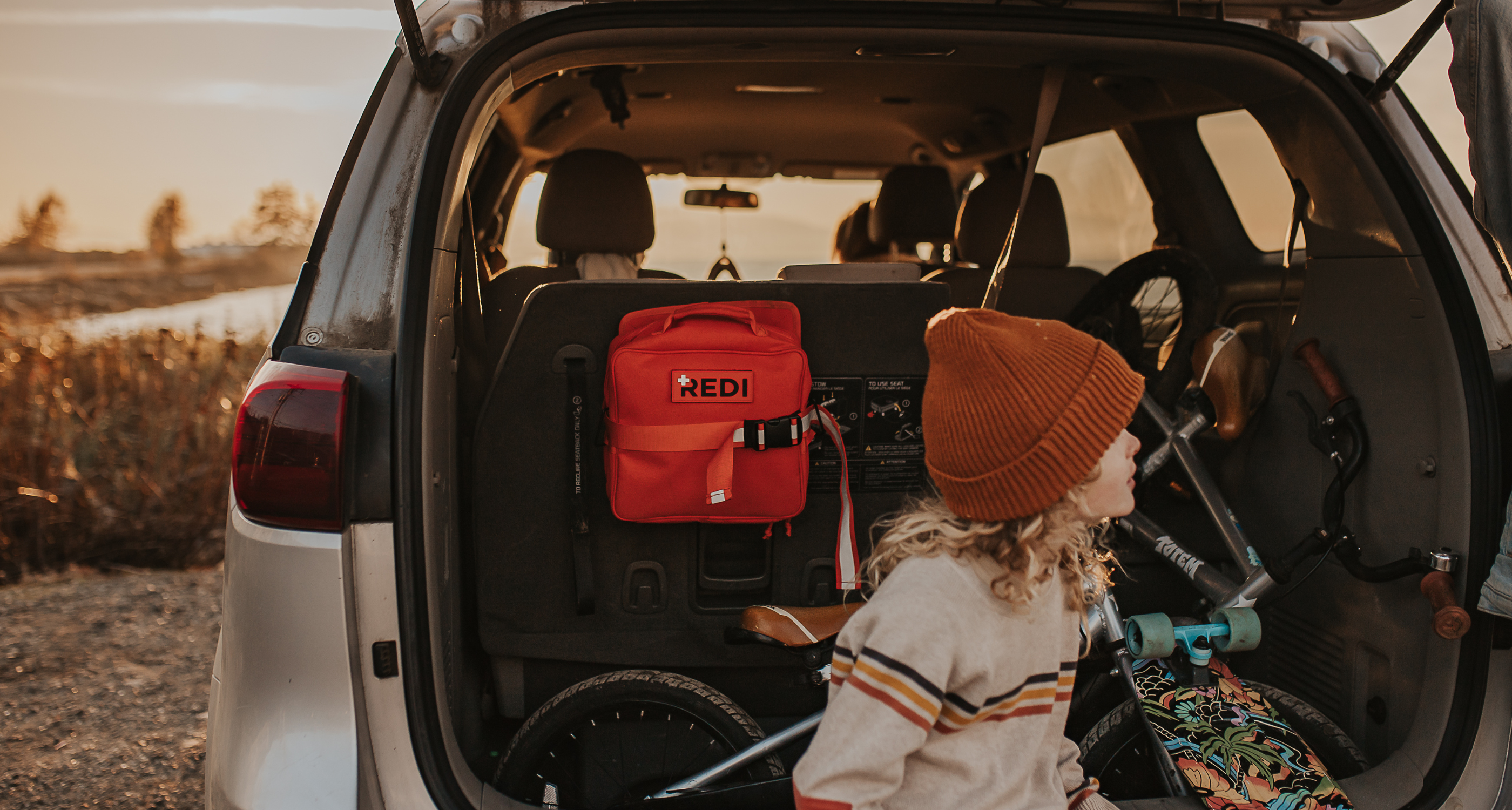 A child sitting on the trunk of a car, with a bike and skateboard in the trunk and the Roadie first aid kit attached to the back of the seat. 