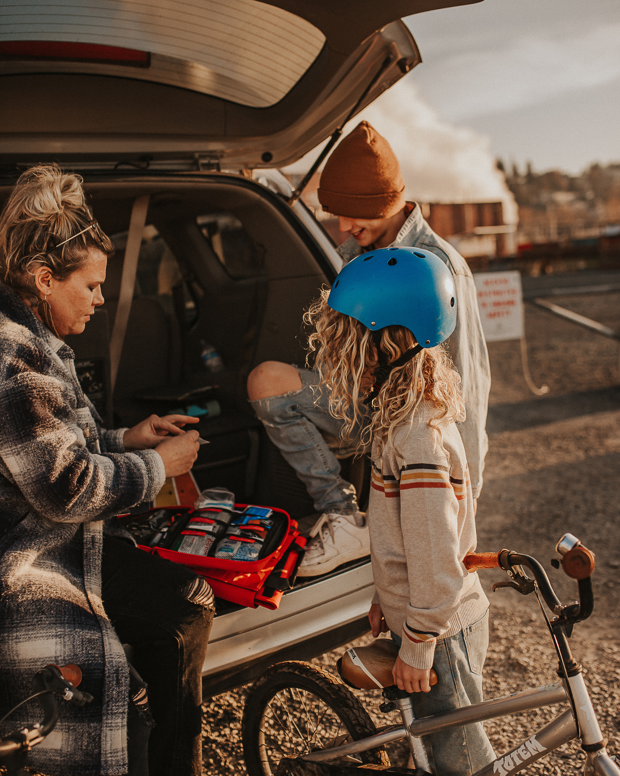 A mother opens the Roadie first aid kit in the trunk of her car at the park, as her two children wait patiently beside her.