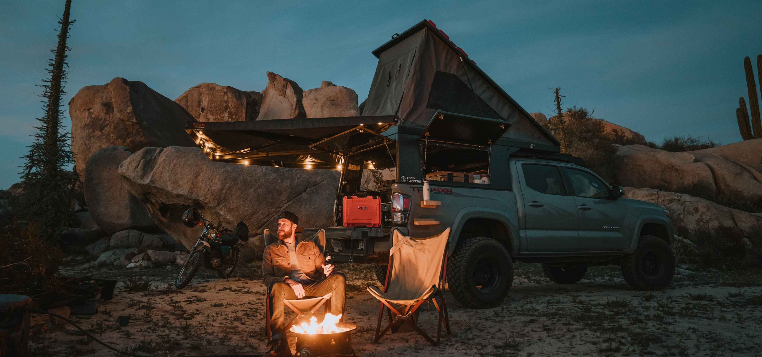 Starry night in the desert as a man sits around a fire with the Roadie Pro + on his truck in the background. 